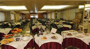 A banquet hall with round tables covered in white tablecloths, each set with plates of food, glasses, and candles. - Park Hotel Cellini (Jesolo)