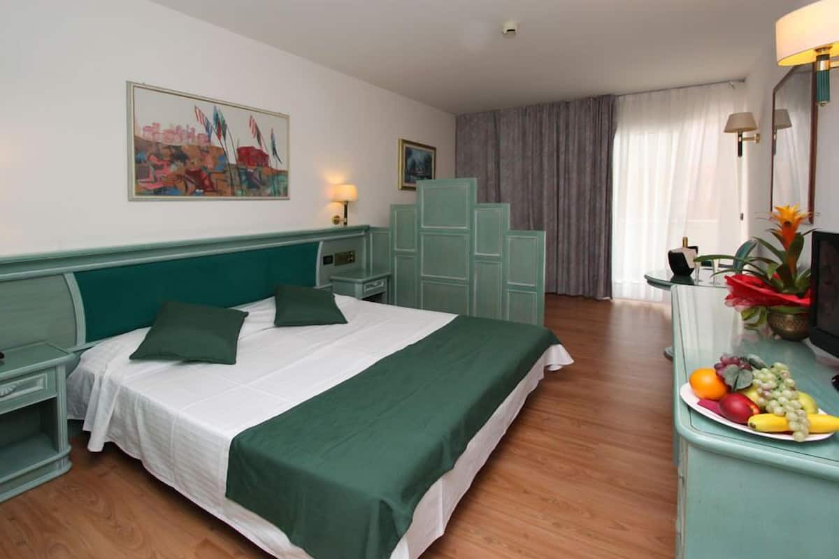 A hotel room with a bed, a desk with a fruit plate, a painting on the wall, and a window with curtains.