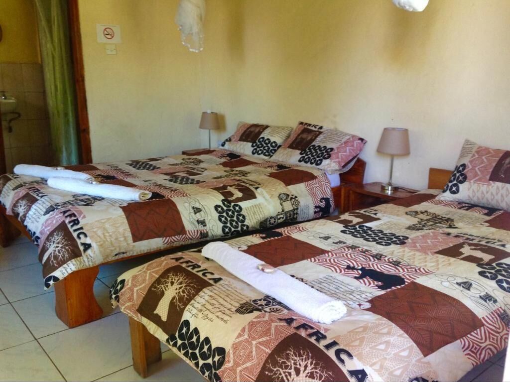 A hotel room with two beds, each covered with a quilt featuring various patterns and the word "AFRICA". There are bedside tables with lamps, and a bathroom is visible in the background.