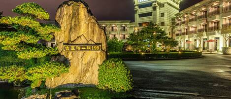 Una entrada de hotel bien iluminada con un gran letrero de piedra, un jardín ajardinado y un edificio de varios pisos con balcones.