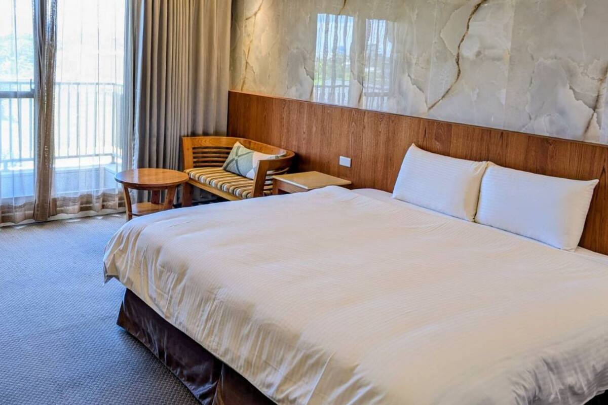 A hotel room with a large bed, a wooden headboard, a small round table, a chair, and a view of the outdoors through large windows.