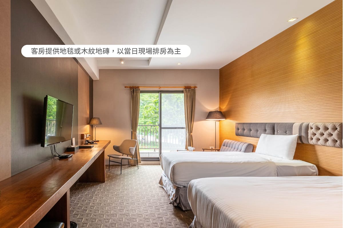 A hotel room with a wooden desk, two beds, a TV, and a view of the outdoors.