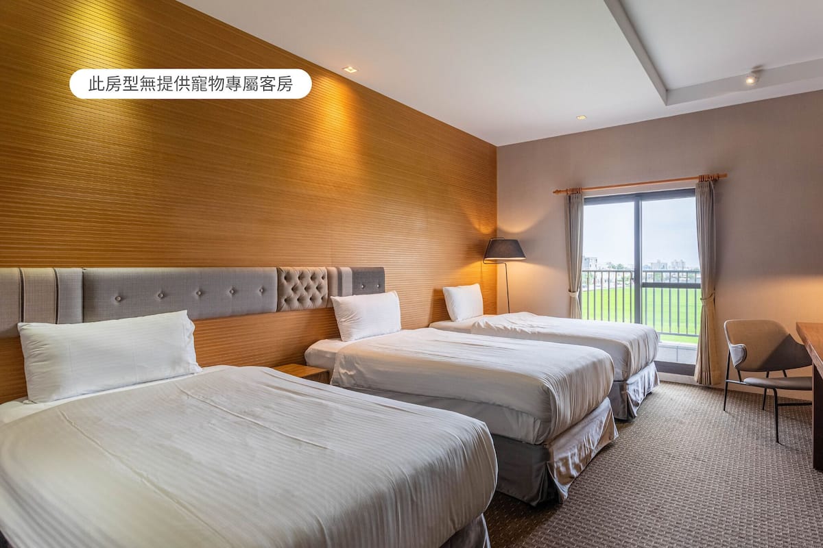 A hotel room with two beds, a desk, and a view of a green landscape.