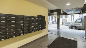 Un área de recepción con una fila de buzones negros etiquetados de B1 a B9, una puerta de cristal de acceso y un vehículo estacionado con la señalización 'RESIDENCE BEREGUNG'.