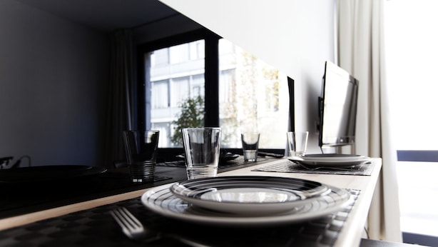 Una mesa de comedor elegantemente dispuesta con platos, vasos y cubiertos, ubicada cerca de una ventana con vistas a edificios.