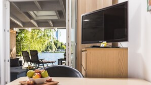 Una habitación moderna con una televisión de pantalla plana sobre un mueble de madera, una mesa de comedor con frutero, y vista a un lago a través de una puerta abierta.