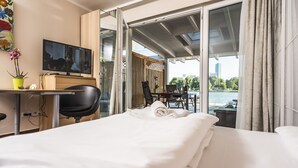 Habitación de hotel con cama, escritorio con televisor, una silla y vista a un cuerpo de agua y paisaje urbano a través de puertas correderas grandes.