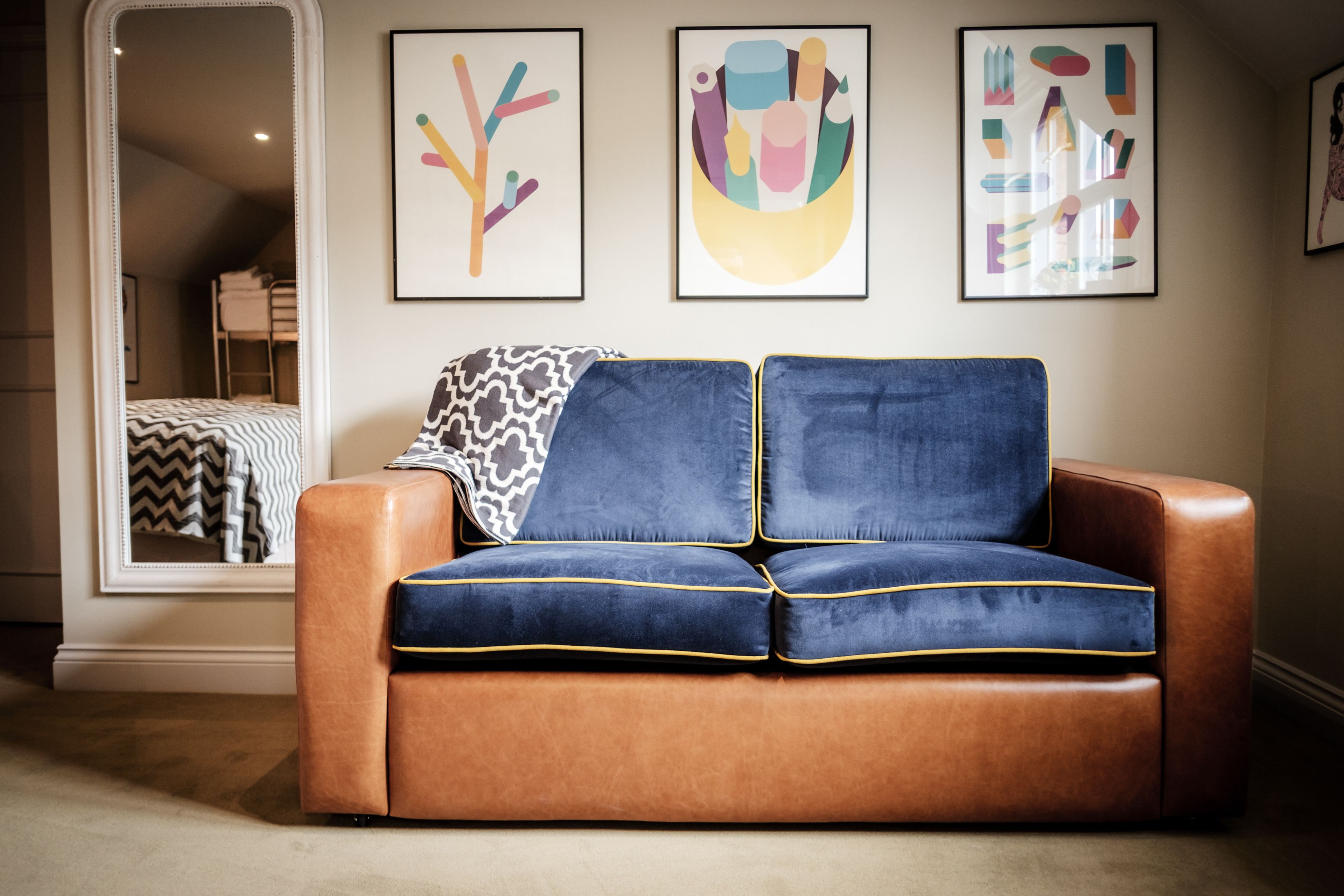 A room with a brown leather sofa, two blue cushions, and a patterned throw. There are abstract paintings on the wall and a mirror reflecting the room.