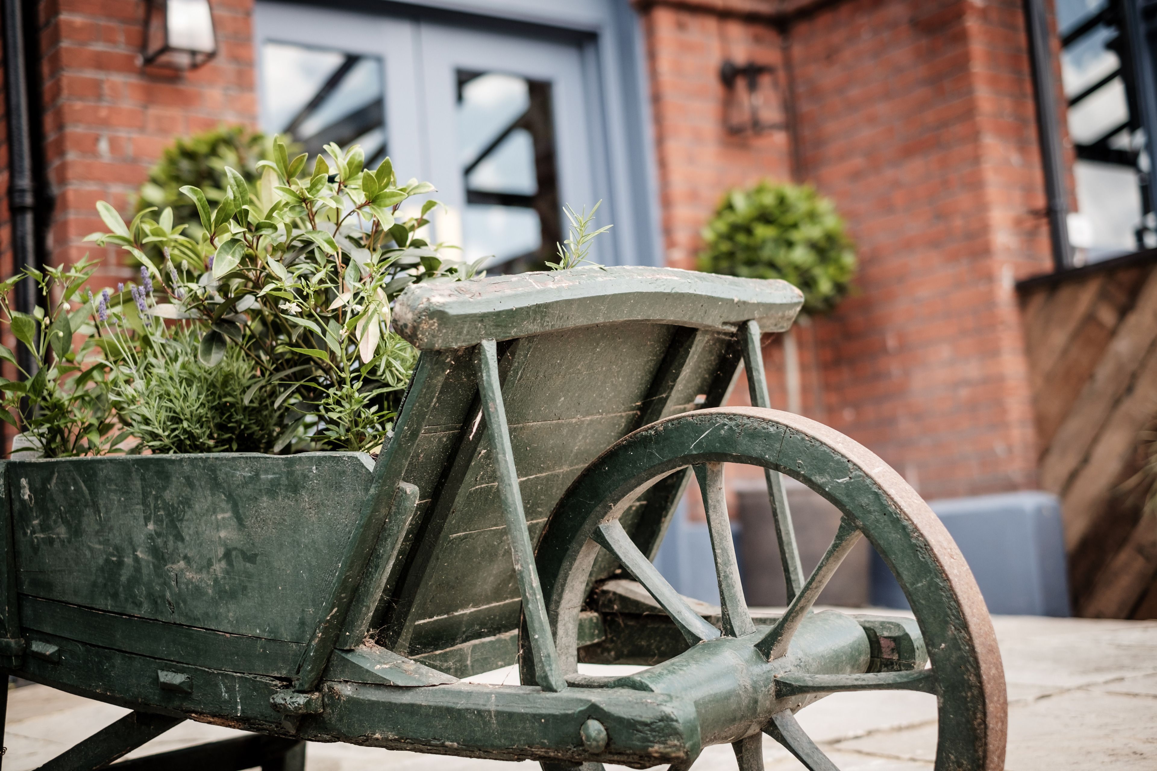 A rustic wooden cart with wheels, positioned in front of a brick building with a blue door and a small potted plant beside it.