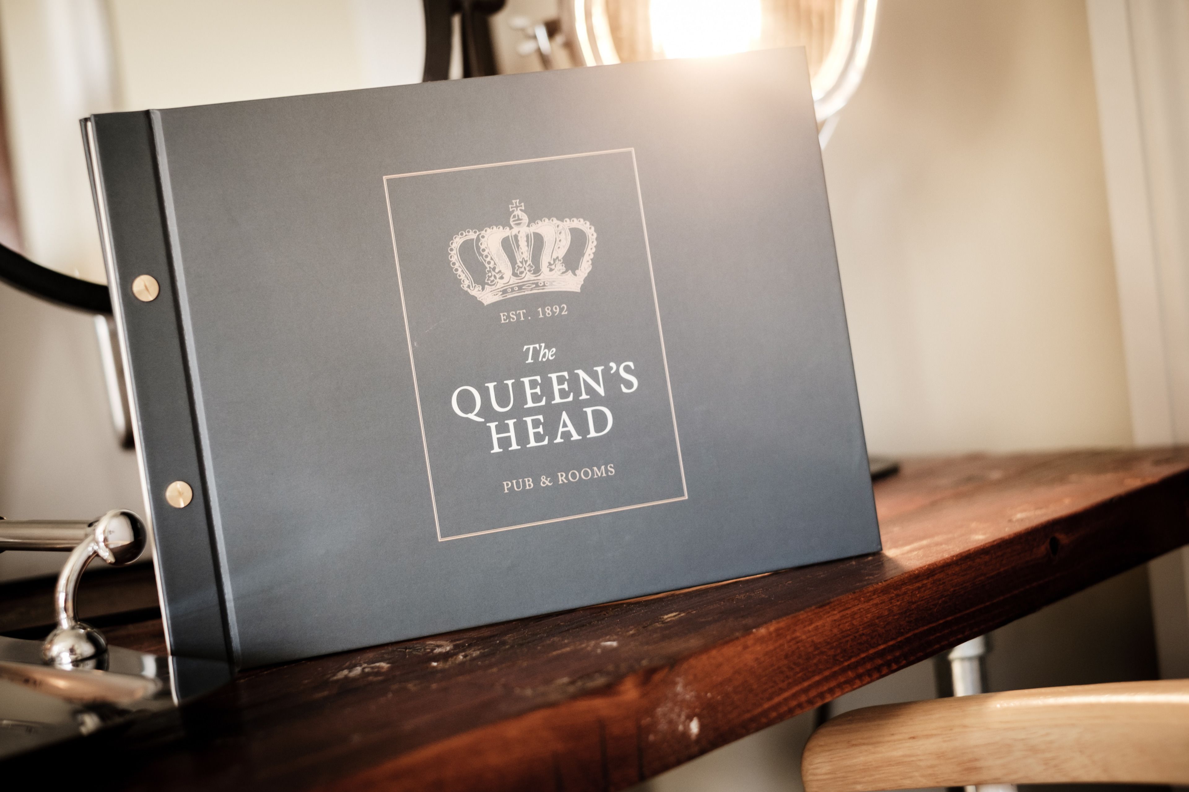 A dark blue folder with a crown emblem and text reading "The Queen's Head PUB & ROOMS" on a wooden surface.