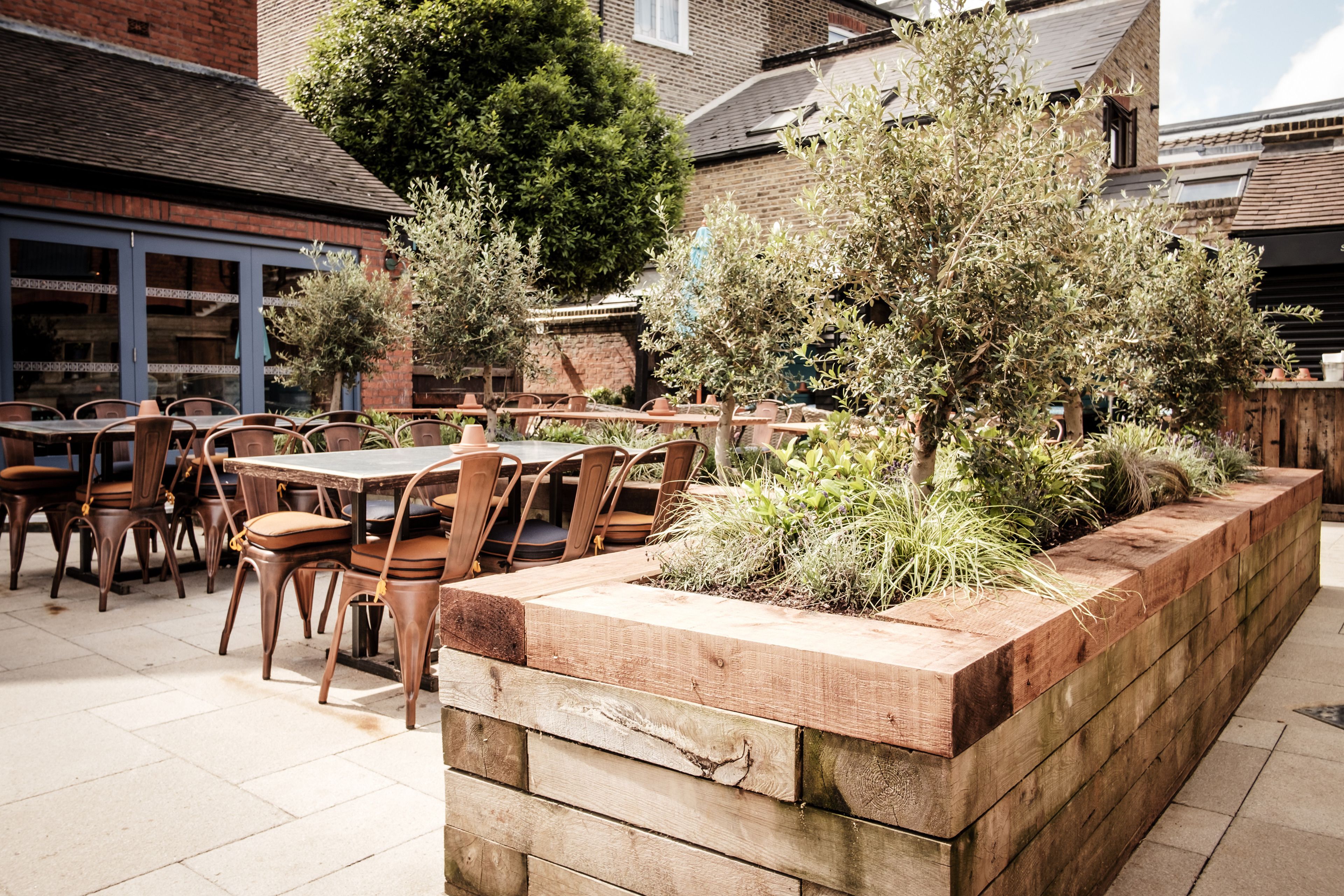An outdoor patio area with wooden tables and chairs, surrounded by a wooden planter with plants.