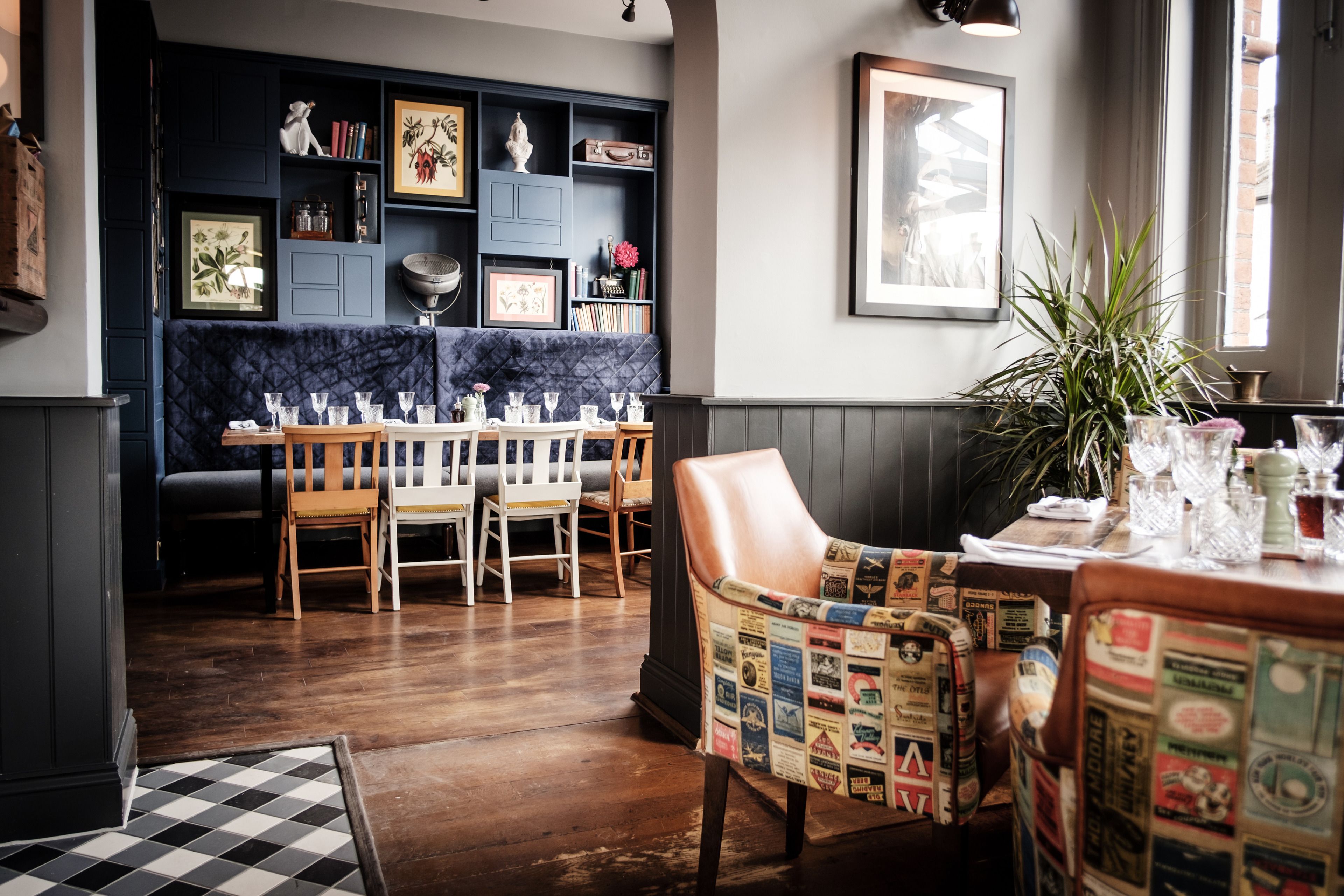 Un restaurante con suelo a cuadros, sillas de madera, una mesa dispuesta con vasos y una pared azul oscuro con estantes.
