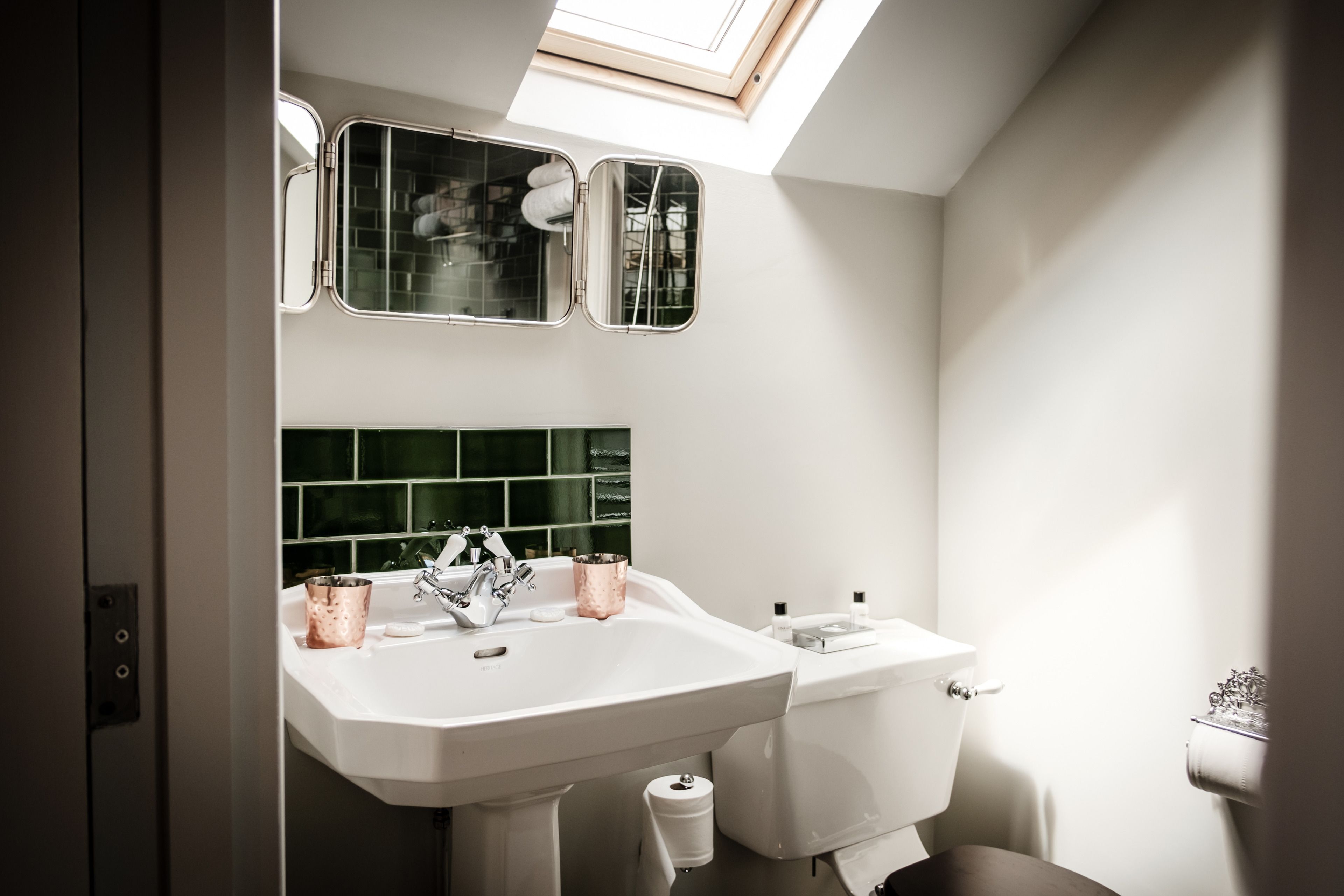 A bathroom with a white sink, two mirrors, a green-tiled backsplash, and a skylight.