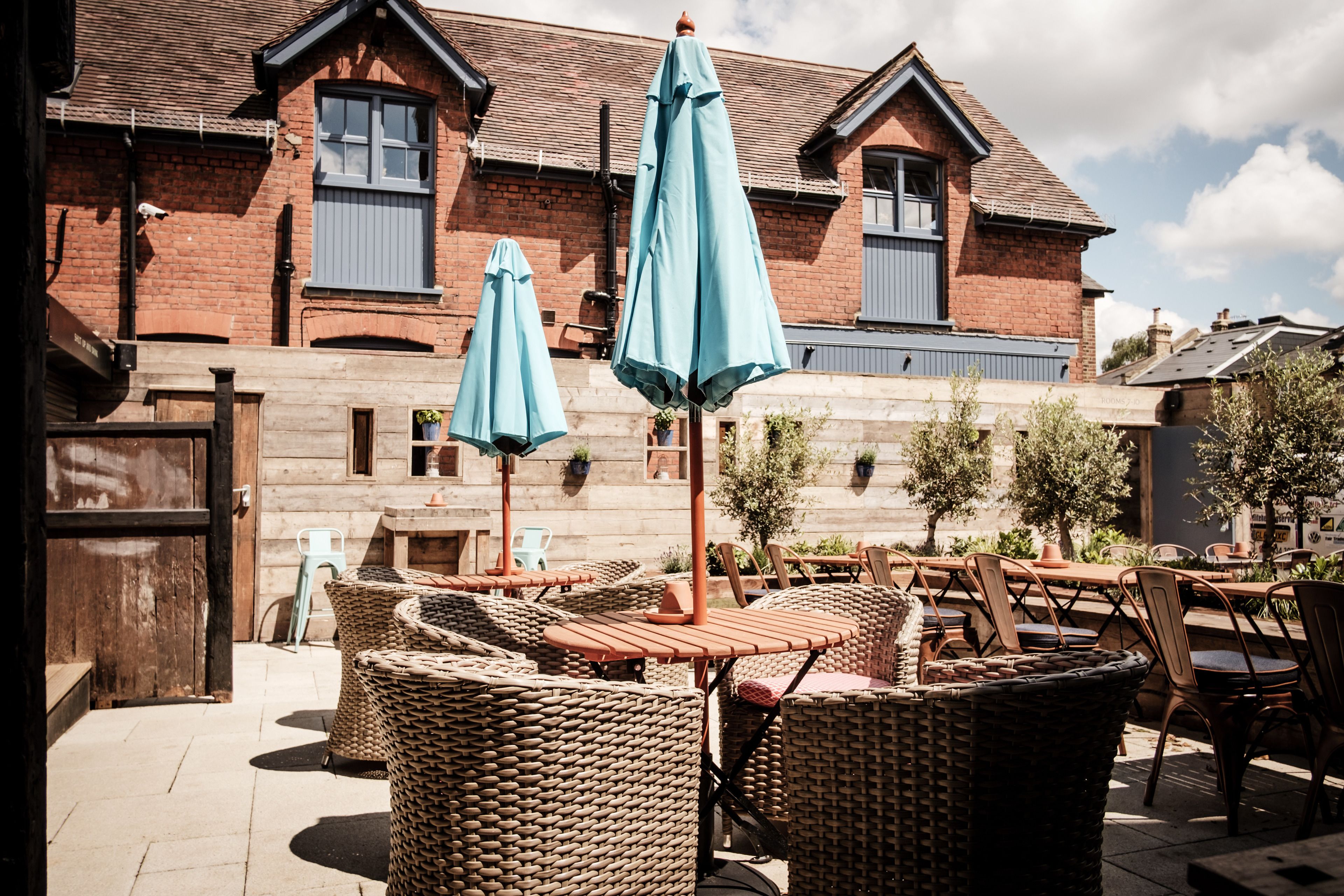 An outdoor seating area with wicker furniture and blue umbrellas, a brick building in the background, and a clear sky.