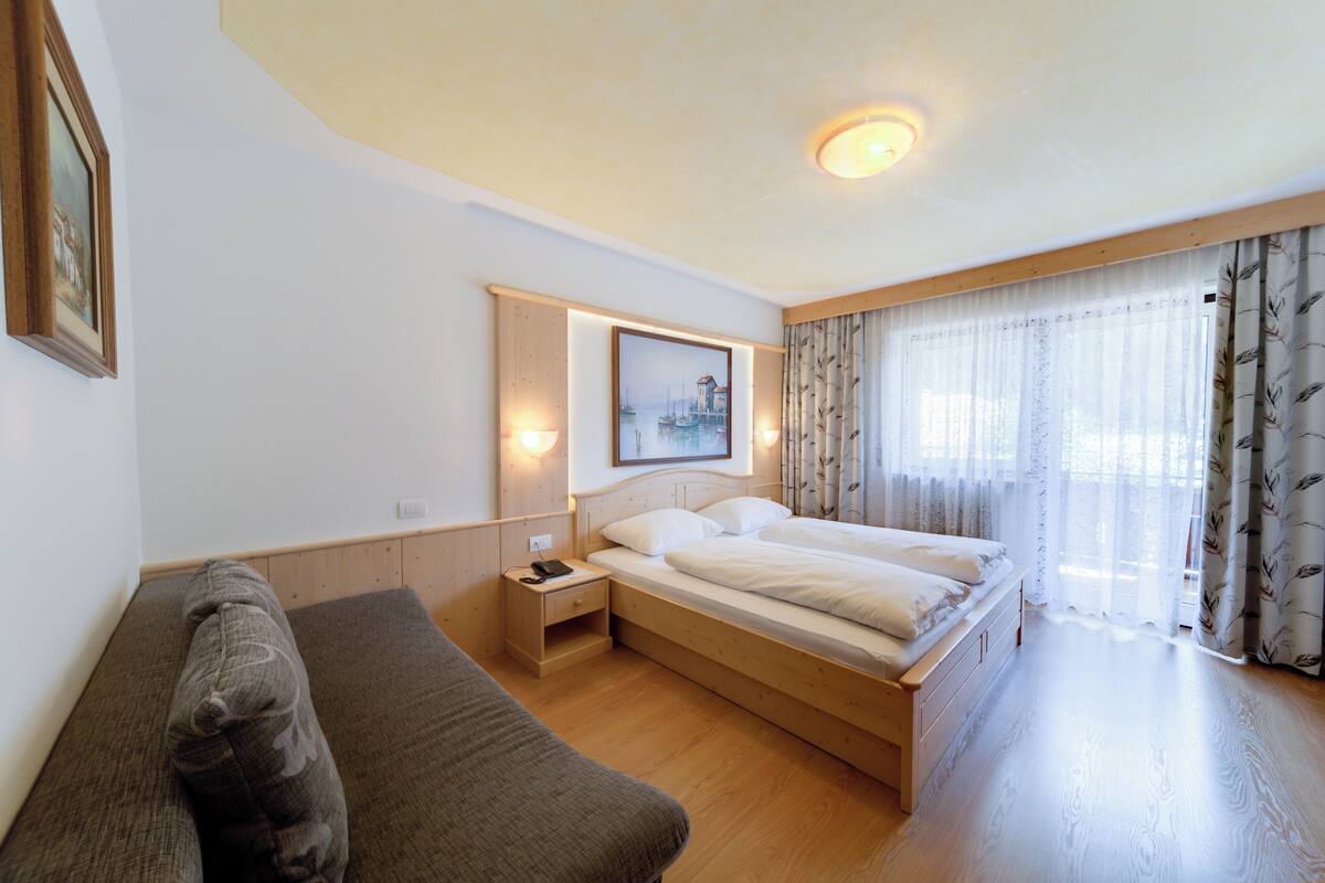 Habitación de hotel con una cama de madera, una mesita de noche, un sofá y un amplio ventanal con cortinas traslúcidas.