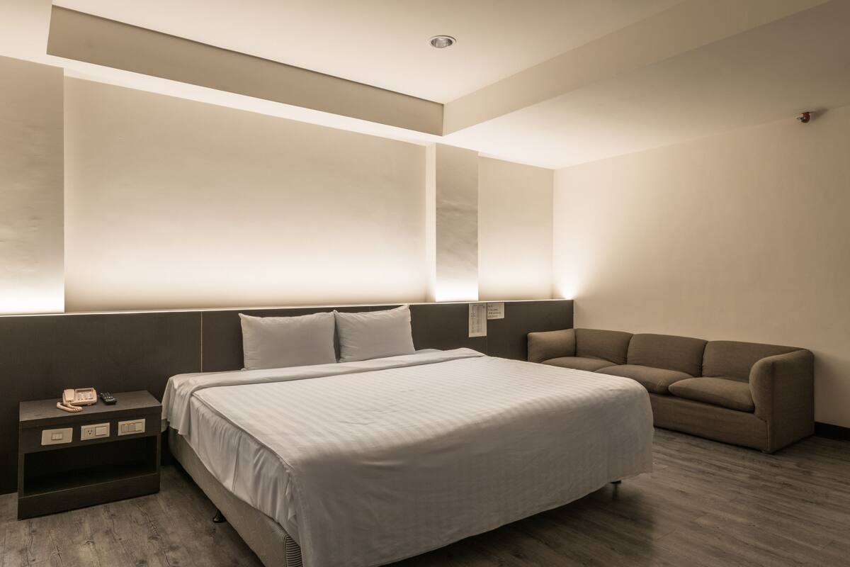 Ein modernes Hotelzimmer mit einem großen Bett, einer Couch, einem Nachttisch mit Telefon und einer wandmontierten Leuchte.