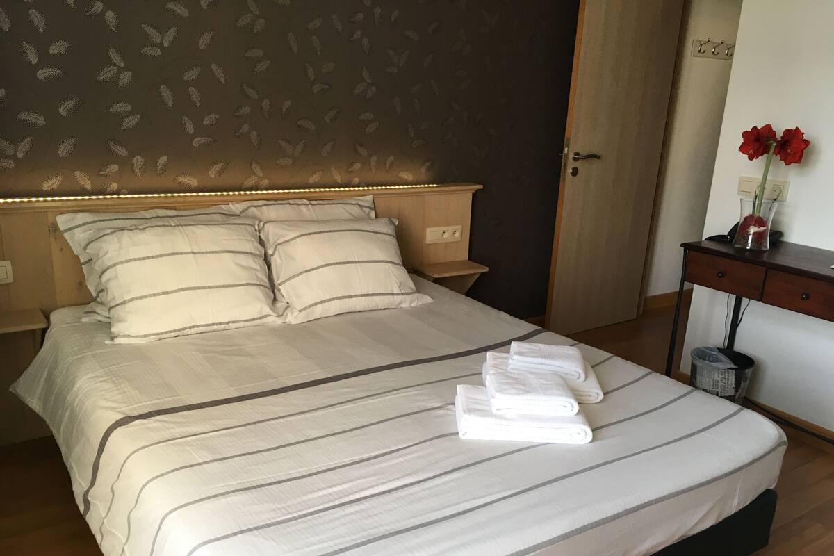 A neatly made bed with striped bedding, a bedside table with a lamp, and a small desk with a vase of red flowers.