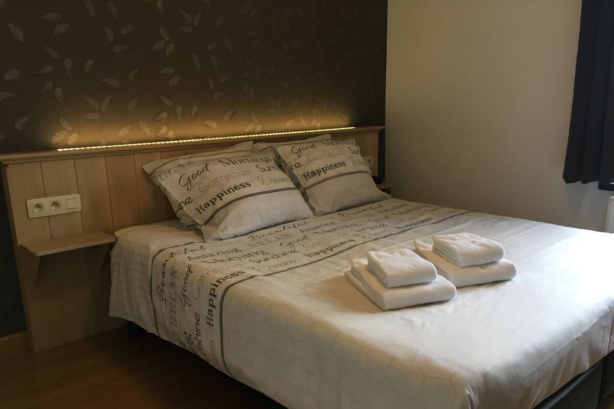 A neatly made bed with a white comforter and two pillows, a bedside table with a lamp, and a wall with a leaf pattern.
