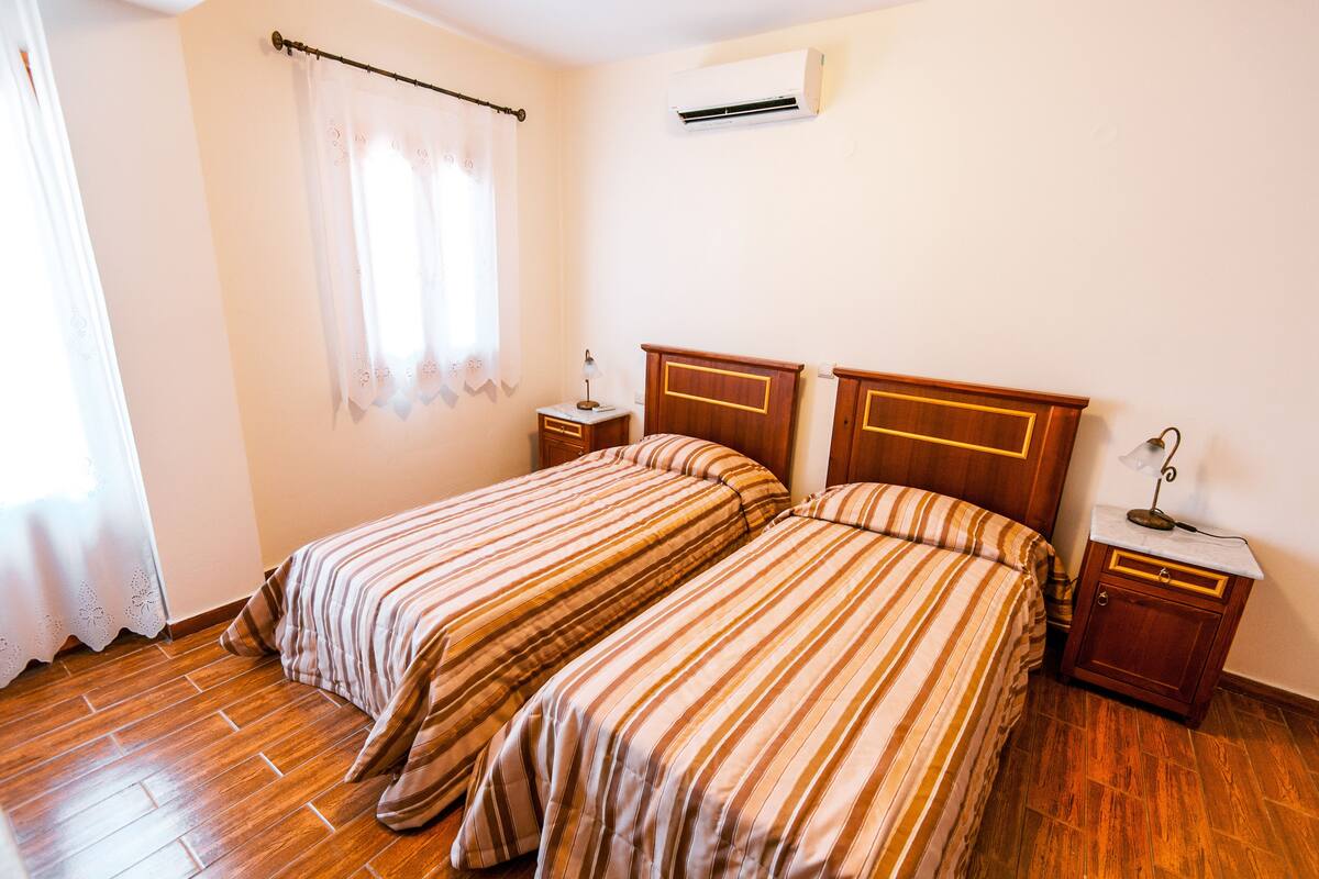 Habitación con dos camas, piso de madera, una mesita de noche con lámpara y un equipo de aire acondicionado en la pared.