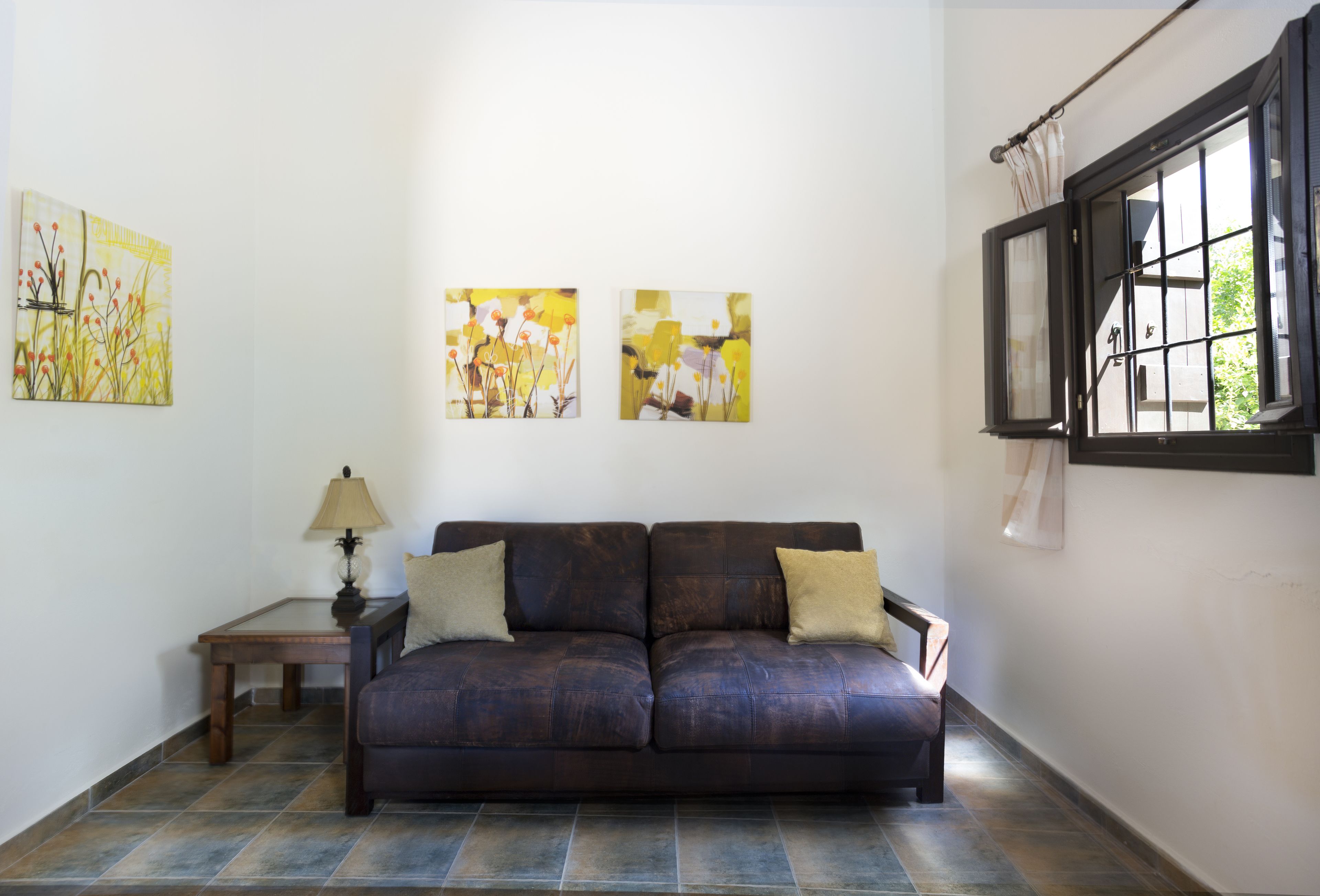 En stue med en mørkebrun lædersofa, et lille træsidebord med en lampe, og to abstrakte billeder på væggen.