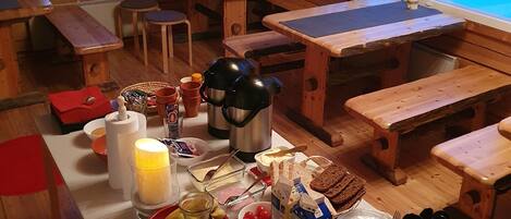 Interior de una cabaña de madera con varias mesas y bancos, un ventanal que ofrece vistas a un paisaje nevado, y una mesa dispuesta con artículos para el desayuno, como pan, huevos y bebidas.