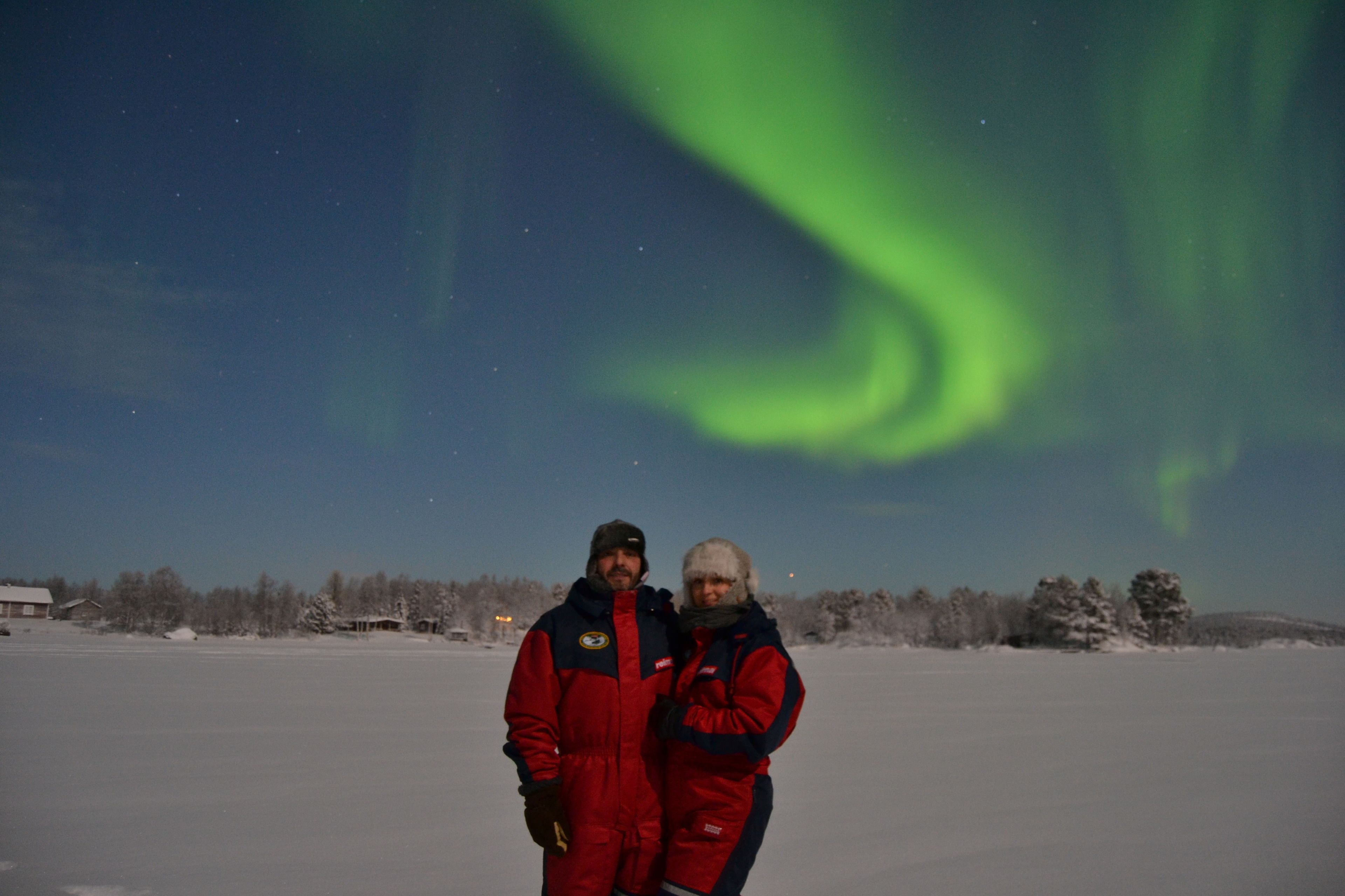 A couple standing on a snow-covered field with the aurora borealis in the background.