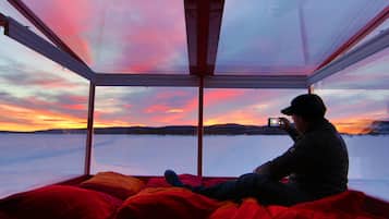 A person sitting on a bed inside a glass-enclosed room with a view of a snowy landscape and a sunset.