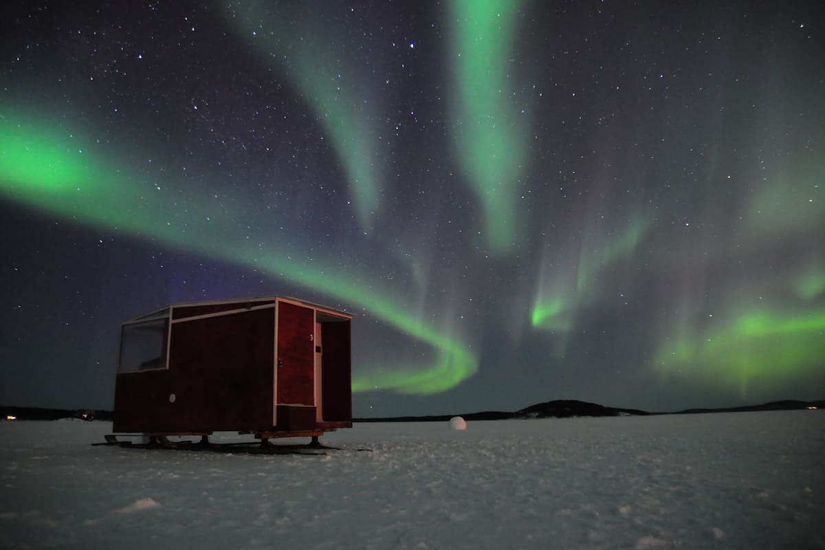 Una pequeña cabaña de madera en un paisaje nevado, con la aurora boreal de fondo.