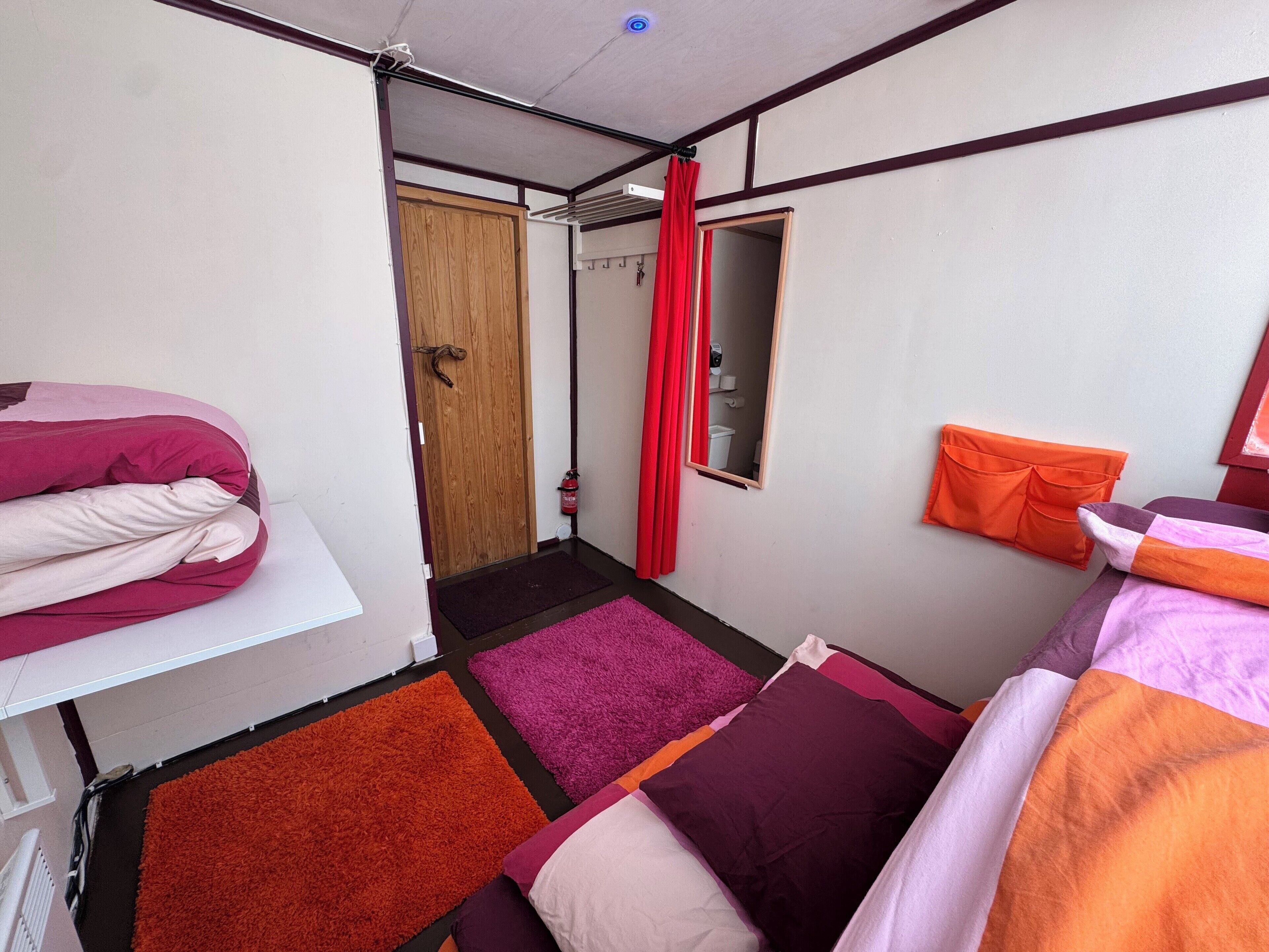 Een knus slaapkamer met een enkel bed, een klein raam met spiegel, een houten deur en een rood gordijn.