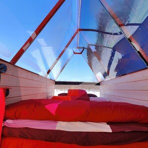 A small, enclosed space with a sloped ceiling and wooden walls, featuring a bed with red bedding and a visible control panel.