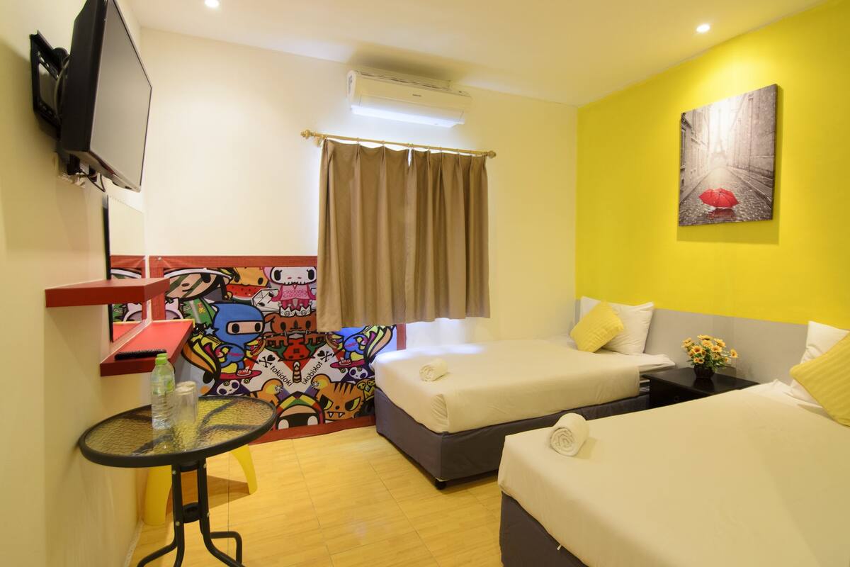 Une chambre d’hôtel avec deux lits, un téléviseur fixé au mur, une petite table avec une bouteille, et une fresque colorée sur le mur.