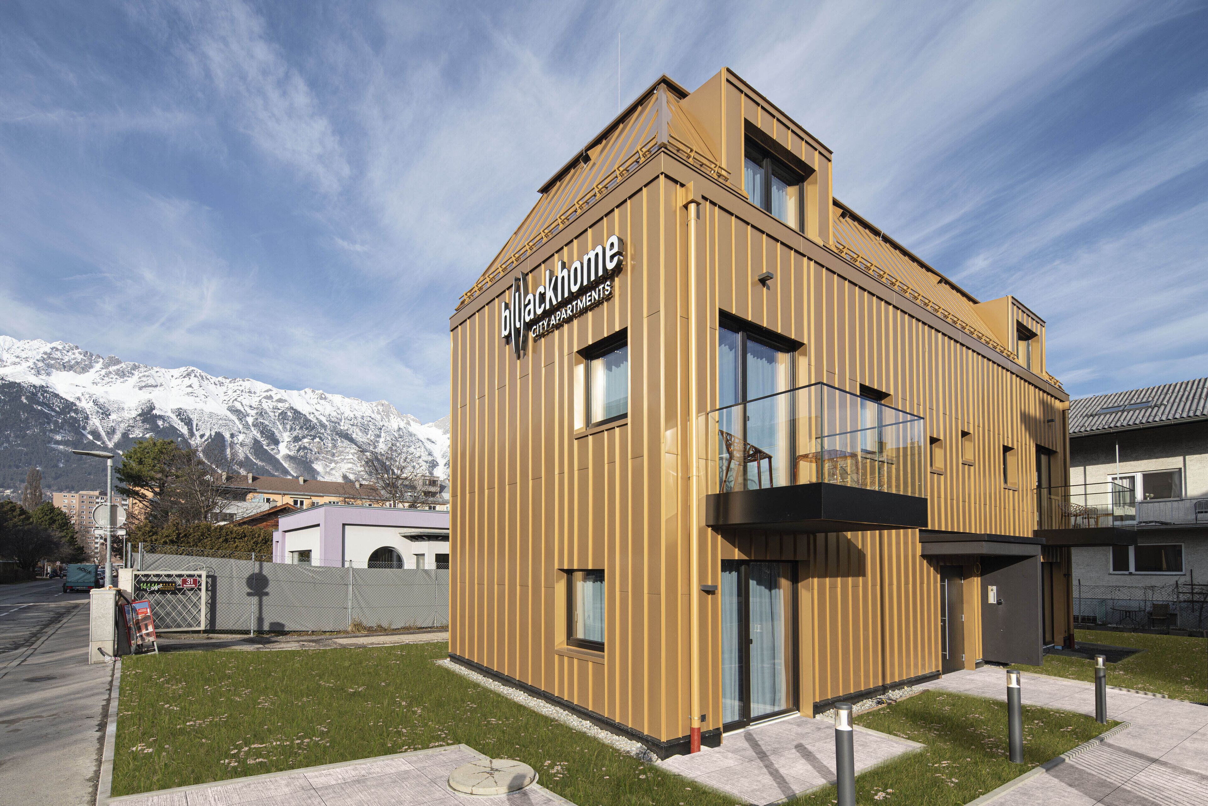A modern building with a sign reading "Blackhome City Apartments" and a view of snow-capped mountains in the background.