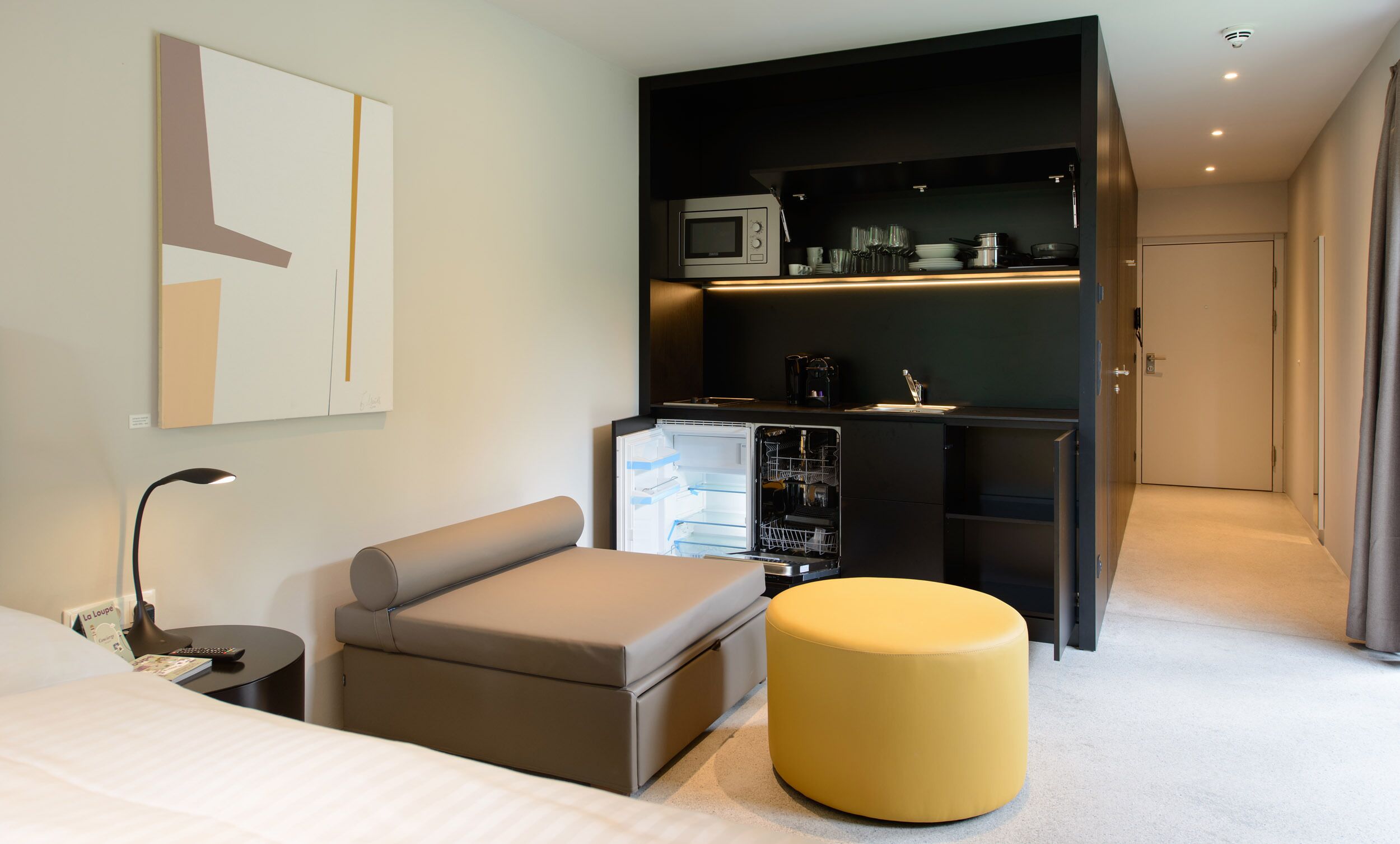A modern hotel room with a bed, a sofa, a small table, a yellow ottoman, a built-in kitchen with a microwave, and a door leading to another room.