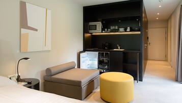 A modern hotel room with a bed, a sofa, a small table, a yellow ottoman, a built-in kitchen with a microwave, and a door leading to another room.