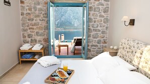Une chambre d’hôtel avec un mur en pierre, un lit avec du linge de maison blanc, un plateau de petit-déjeuner et une vue sur un balcon avec une table et des chaises.
