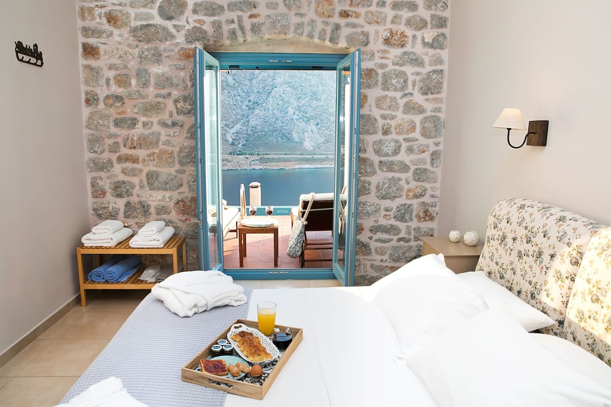 Hotelkamer met een stenen muur, een bed met witte lakens, een ontbijttafel en uitzicht op een balkon met een tafel en stoelen.