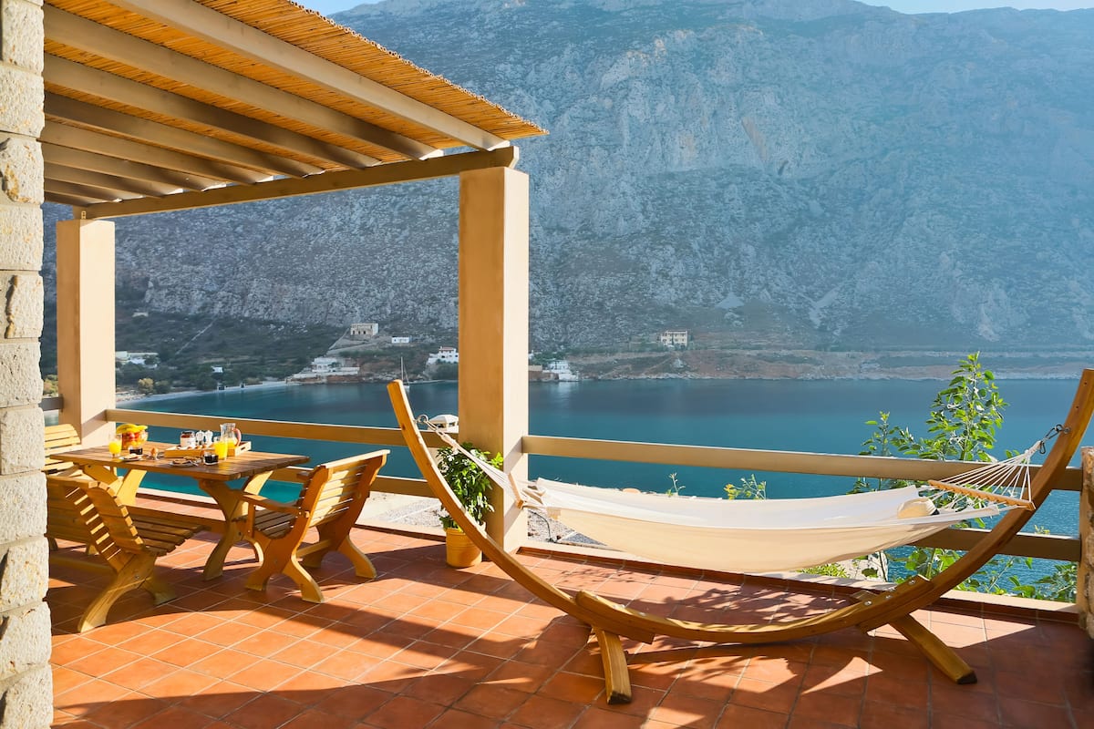 Een terras met een houten tafel en stoelen, een hangmat, en uitzicht op een meer en bergen.