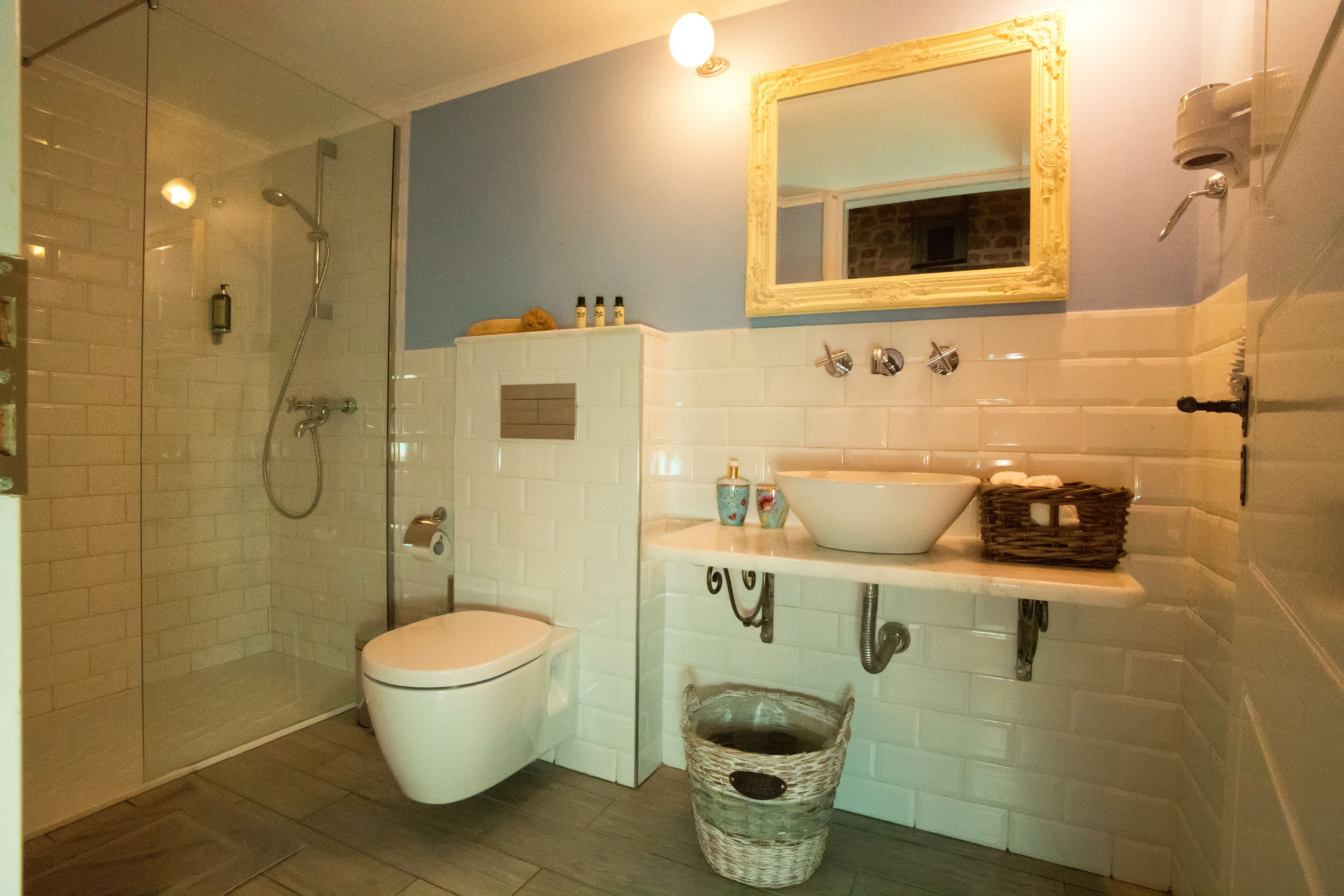 A bathroom with a glass-enclosed shower, a toilet, a sink with a mirror above it, and a basket on the floor.