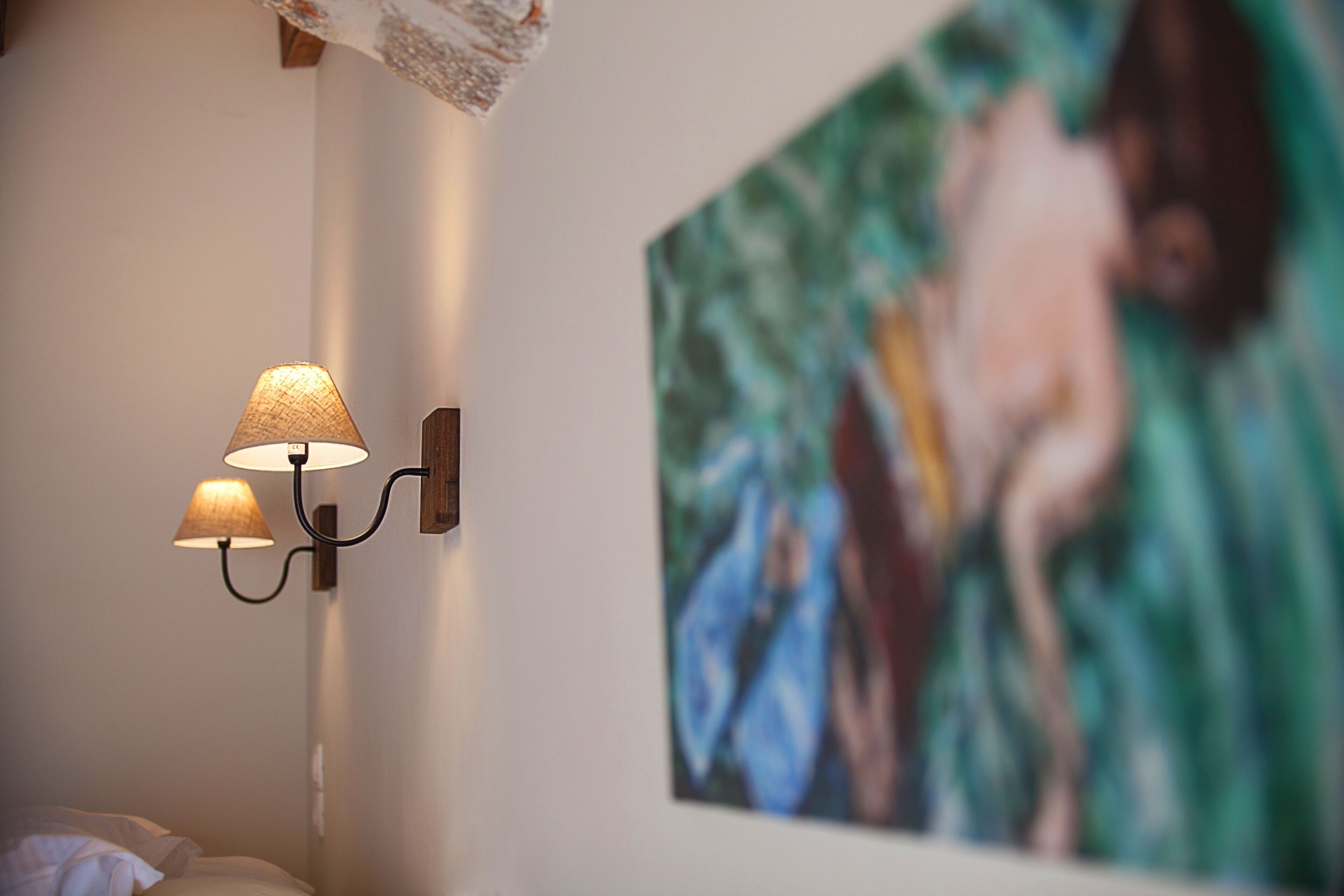En vägglampa med två lampskärmar, en målning som föreställer en person i en blå klänning och en säng med vita sängkläder.