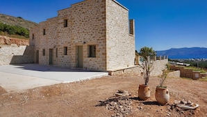 Un bâtiment en pierre doté d’un toit plat, d’une zone pavée à l’avant et offrant une vue sur des montagnes lointaines.