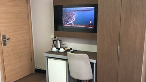 Quarto de hotel com chão de madeira, uma escrivaninha com cadeira, televisão na parede e um guarda-roupa.