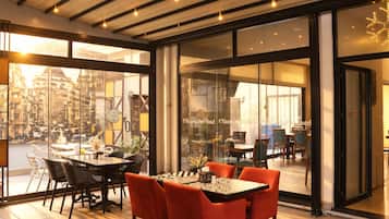 Un intérieur d’hôtel moderne avec un espace repas comprenant une table et des chaises, de grandes fenêtres offrant une vue sur la ville et une cloison en verre portant le nom de l’hôtel.