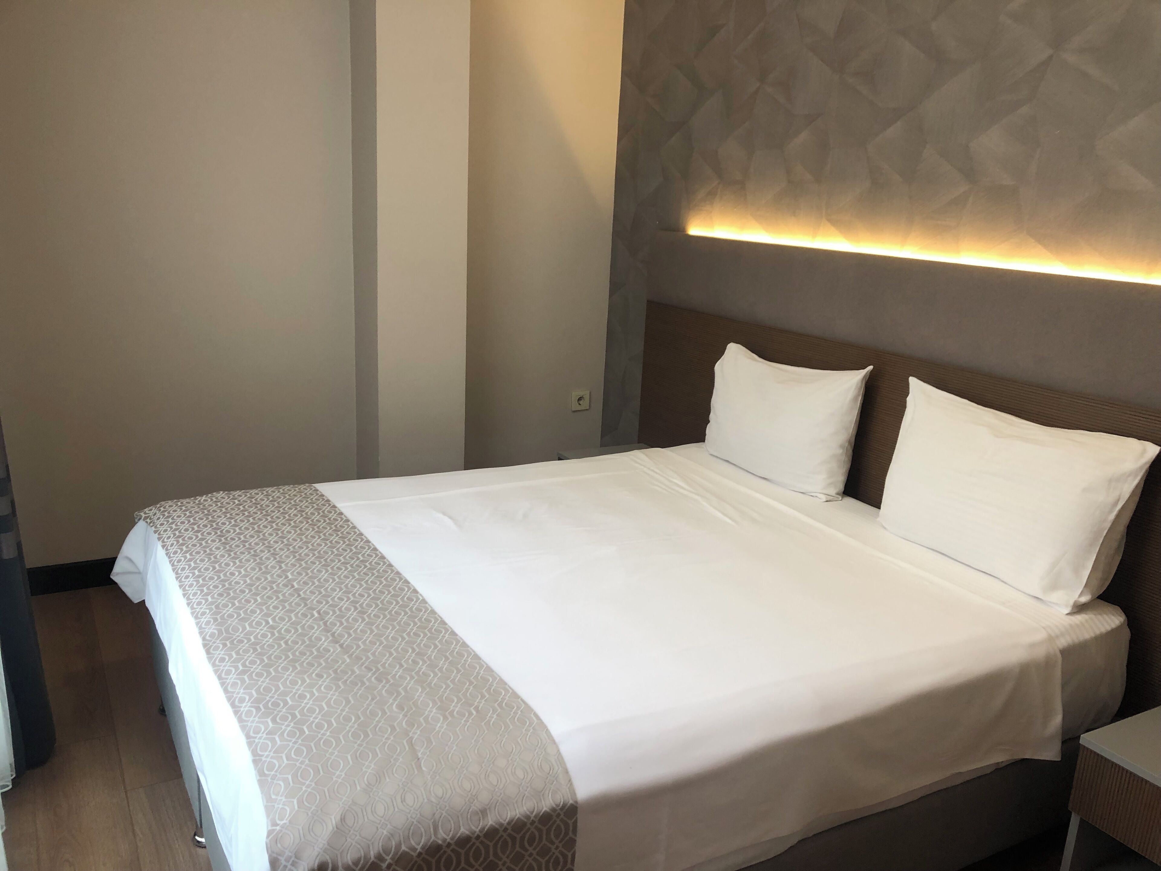 A neatly made bed with a patterned bedspread and a white throw blanket, a wooden headboard, and a wall-mounted light fixture.