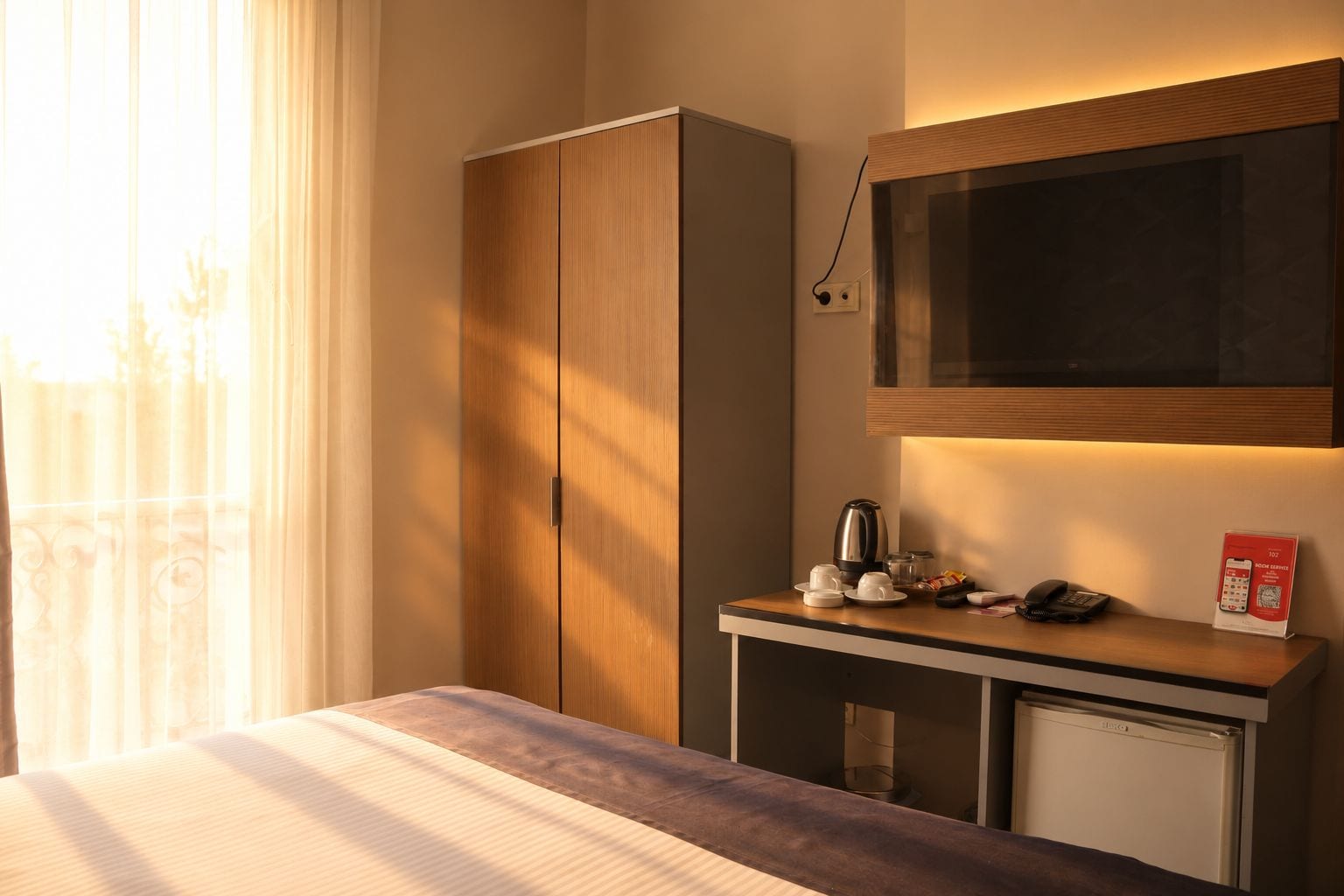 A hotel room with a bed, a wooden wardrobe, a desk with a kettle, a phone, and a TV.