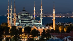 Une mosquée historique, dotée de plusieurs minarets et de coupoles, entourée d’arbres et surplombant une étendue d’eau.