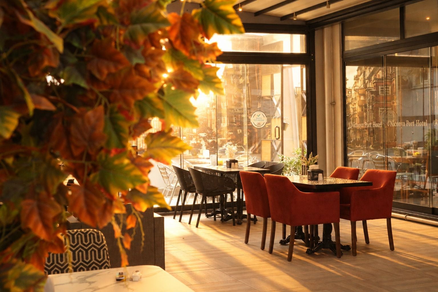 An indoor dining area with tables and chairs, a glass wall with a view of the city, and a decorative plant in the foreground.