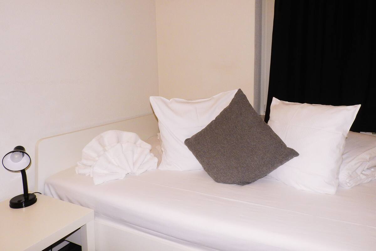 A neatly made bed with white and gray pillows, a white bedside table with a lamp, and a black curtain partially drawn.