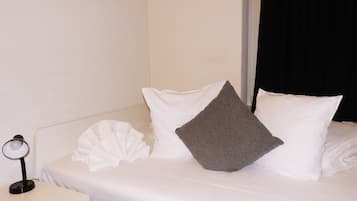 Una cama bien hecha con almohadas blancas y grises, una mesita de noche blanca con una lámpara y una cortina negra parcialmente abierta.