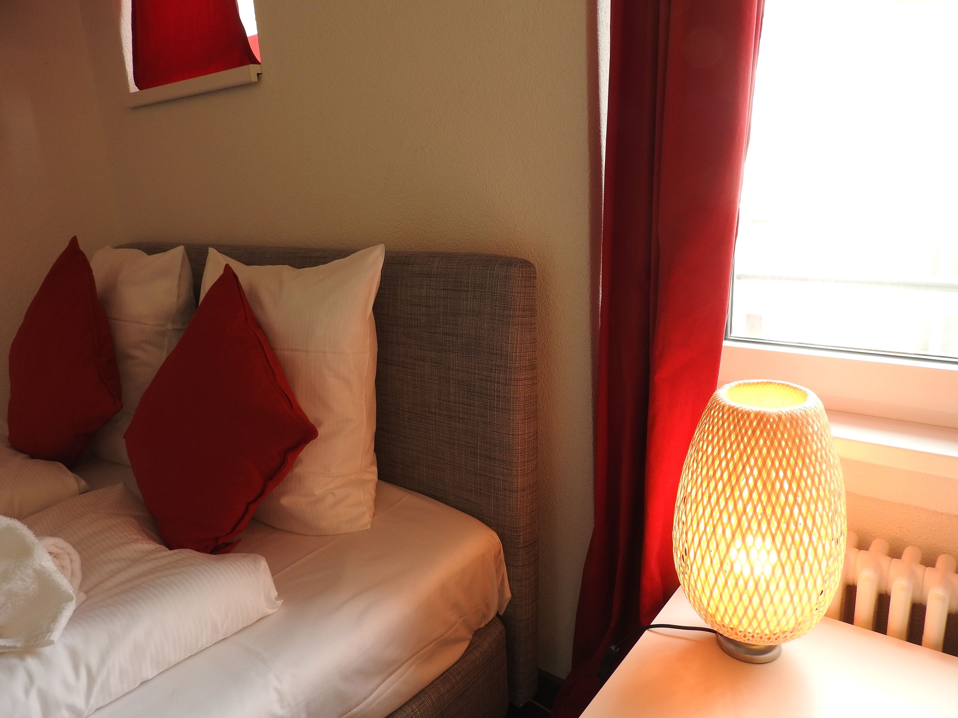 Een bed met witte en rode kussens, een grijze hoofdbord en een lamp op een nachtkastje.