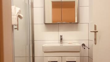 Une salle de bain avec une douche à parois de verre, un lavabo blanc avec un miroir au-dessus et une petite poubelle.